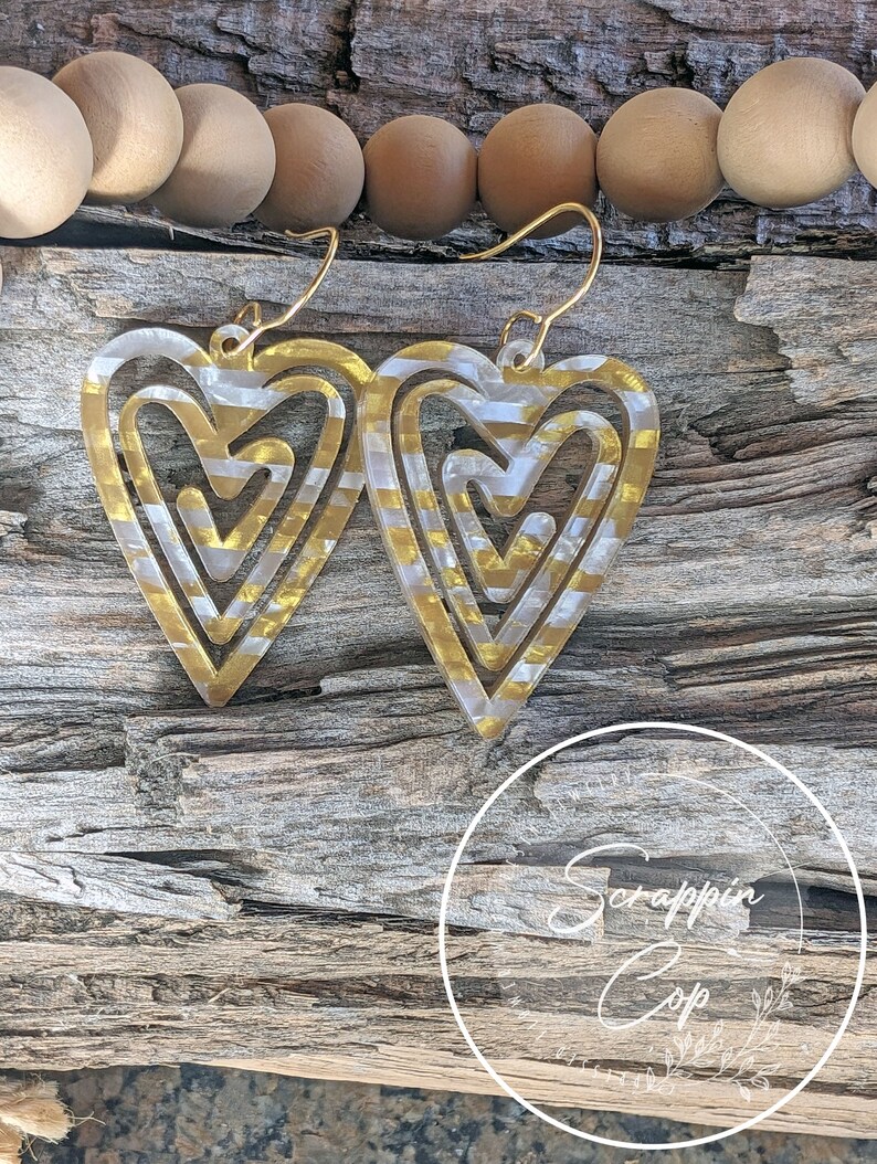 Puede incluir: Pendientes de coraz&oacute;n en capas de oro y blanco. Los corazones est&aacute;n superpuestos y tienen un patr&oacute;n de espiral. Los pendientes cuelgan de ganchos dorados.