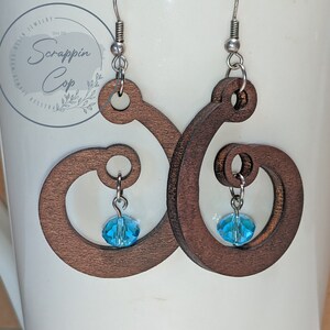 Pode incluir: Um par de brincos de madeira castanho com um design em espiral. Cada brinco tem um pequeno cristal azul pendurado.
