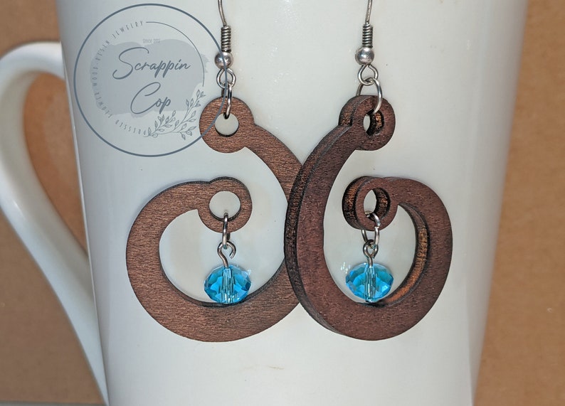 Pode incluir: Um par de brincos de madeira castanho com um design em espiral. Cada brinco tem um pequeno cristal azul pendurado.