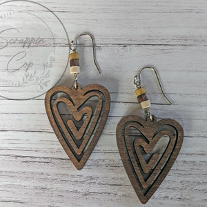 Puede incluir: Un par de pendientes de madera en forma de coraz&oacute;n con un dise&ntilde;o en capas. Los pendientes son marrones y tienen un gancho plateado. Cada pendiente tiene tres cuentas de madera en marr&oacute;n, beige y amarillo.