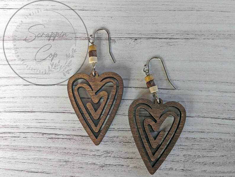 Puede incluir: Un par de pendientes de madera en forma de coraz&oacute;n con un dise&ntilde;o en capas. Los pendientes son marrones y tienen un gancho plateado. Cada pendiente tiene tres cuentas de madera en marr&oacute;n, beige y amarillo.