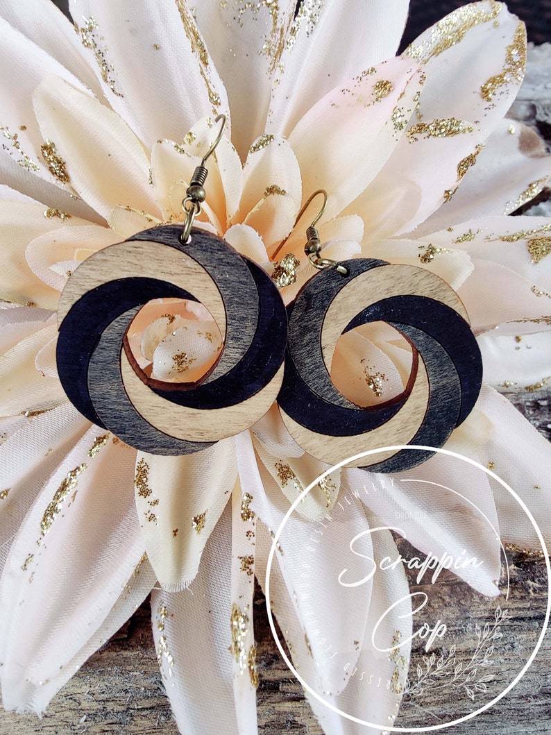 May include: A pair of wooden earrings with a black and natural wood swirl design. The earrings are hanging from gold-toned hooks.