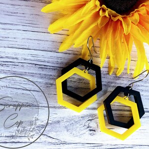 May include: A pair of black and yellow hexagon shaped earrings with a silver hook. The earrings are layered with a smaller hexagon inside a larger hexagon.