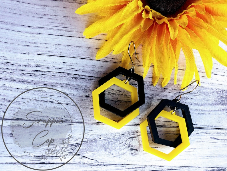 May include: A pair of black and yellow hexagon shaped earrings with a silver hook. The earrings are layered with a smaller hexagon inside a larger hexagon.