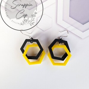 May include: A pair of yellow and black hexagon shaped earrings. The earrings are made of acrylic and have a geometric design.