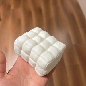 May include: A small, rectangular, white foam block featuring a grid of raised, rounded, quilted-style squares on its top surface. The object has a soft, matte texture and a uniform white color throughout its entire visible structure.