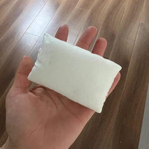May include: A small, rectangular, white pillow with a smooth, solid-colored surface. The object is shaped like a miniature cushion and features clean, stitched edges along its perimeter, presenting a simple and uniform appearance throughout.