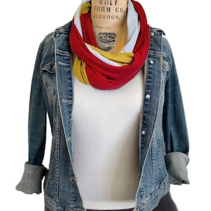 K&ouml;nnte beinhalten: Jeansjacke mit hochgekrempelten &Auml;rmeln &uuml;ber einem wei&szlig;en Ripp-Tanktop. Ein mehrfarbiger Infinity-Schal in Rot, Wei&szlig; und Gold ist um den Hals der Schaufensterpuppe drapiert. Die Schaufensterpuppe steht vor wei&szlig;em Hintergrund.