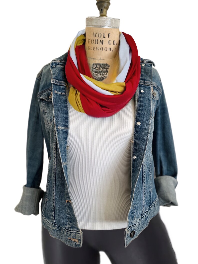 K&ouml;nnte beinhalten: Jeansjacke mit hochgekrempelten &Auml;rmeln &uuml;ber einem wei&szlig;en Ripp-Tanktop. Ein mehrfarbiger Infinity-Schal in Rot, Wei&szlig; und Gold ist um den Hals der Schaufensterpuppe drapiert. Die Schaufensterpuppe steht vor wei&szlig;em Hintergrund.