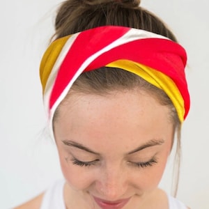 K&ouml;nnte beinhalten: Ein farbenfrohes Stirnband mit einem rot, wei&szlig; und gelb gestreiften Design. Das Stirnband ist um den Kopf gewickelt, wobei der obere Teil des Kopfes und die Stirn sichtbar sind. Das Haar ist zu einem Dutt gebunden.