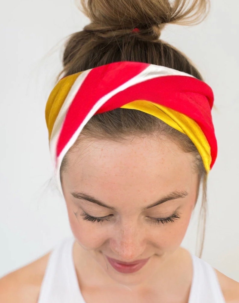 K&ouml;nnte beinhalten: Ein farbenfrohes Stirnband mit einem rot, wei&szlig; und gelb gestreiften Design. Das Stirnband ist um den Kopf gewickelt, wobei der obere Teil des Kopfes und die Stirn sichtbar sind. Das Haar ist zu einem Dutt gebunden.