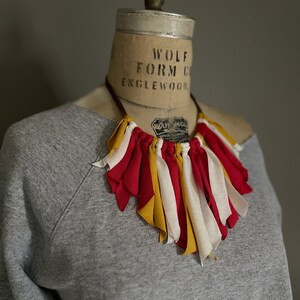 K&ouml;nnte beinhalten: Eine handgefertigte Halskette mit roten, gelben und wei&szlig;en Stoffstreifen. Die Halskette wird auf einer Vintage-Schneiderpuppe mit einem grauen Sweatshirt pr&auml;sentiert. Ein einzigartiges, farbenfrohes Schmuckst&uuml;ck.