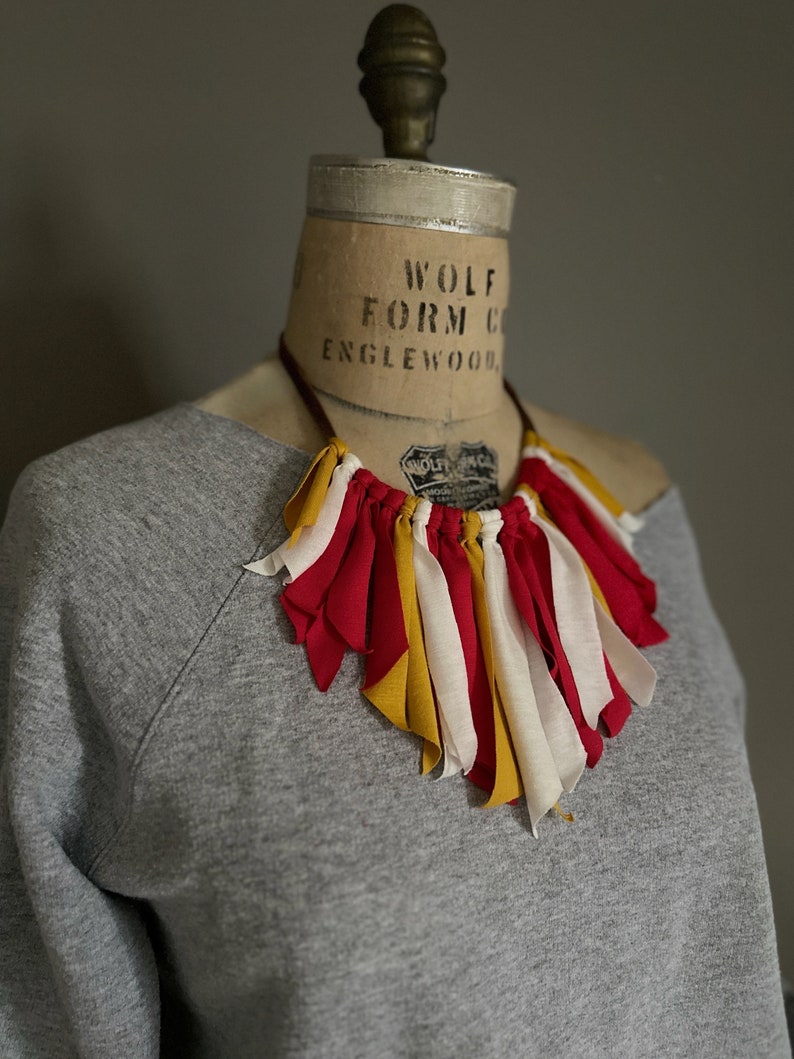 K&ouml;nnte beinhalten: Eine handgefertigte Halskette mit roten, gelben und wei&szlig;en Stoffstreifen. Die Halskette wird auf einer Vintage-Schneiderpuppe mit einem grauen Sweatshirt pr&auml;sentiert. Ein einzigartiges, farbenfrohes Schmuckst&uuml;ck.