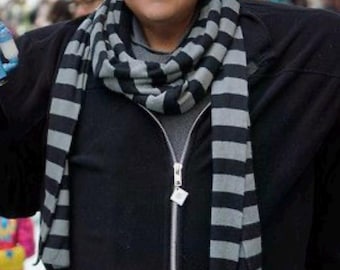 May include: A person dressed in a costume featuring a black zip-up jacket, a grey shirt, and a long scarf with horizontal black and grey stripes. The individual has a bald head, prominent dark eyebrows, and a distinctively shaped nose.