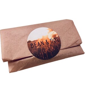 May include: A rectangular package wrapped in brown paper. A round sticker with the words "BEAN & DOT" is affixed to the package. The sticker features a sunset scene with a field of tall grass.