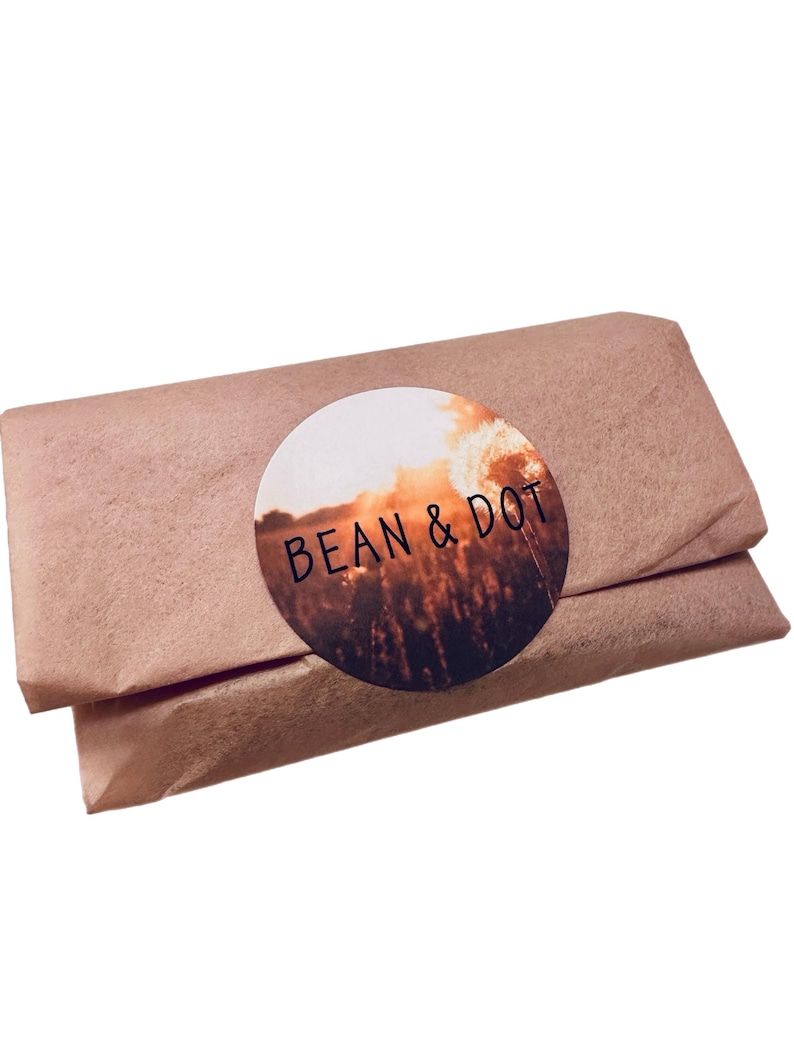 May include: A rectangular package wrapped in brown paper. A round sticker with the words "BEAN & DOT" is affixed to the package. The sticker features a sunset scene with a field of tall grass.