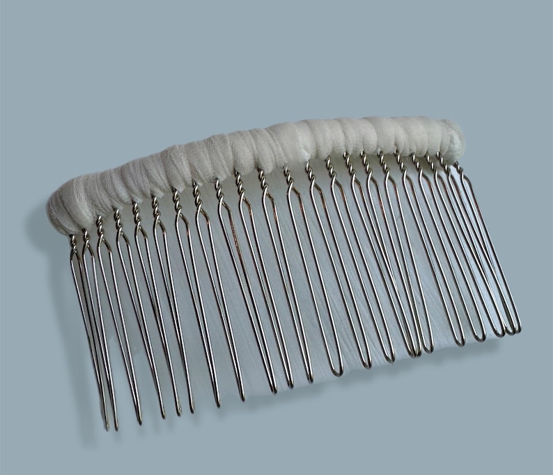 May include: A bridal hair comb with a row of silver-colored metal teeth attached to a white fabric base. The comb's teeth are evenly spaced and slightly curved. The fabric base is a soft, off-white color.