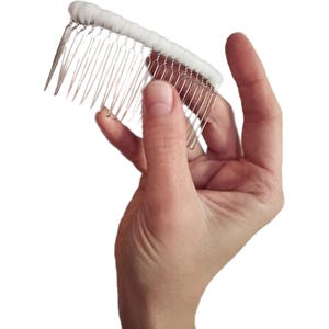 May include: A white bridal hair comb with silver metal teeth. The comb features a row of white fabric along the top, designed to hold a veil or add a decorative element to a hairstyle. The comb is held in a hand.