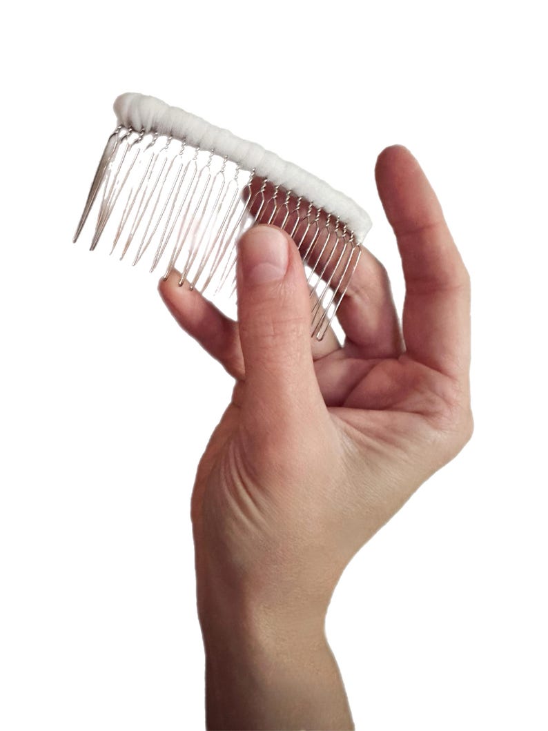 May include: A white bridal hair comb with silver metal teeth. The comb features a row of white fabric along the top, designed to hold a veil or add a decorative element to a hairstyle. The comb is held in a hand.