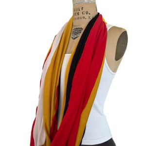 K&ouml;nnte beinhalten: Ein mehrfarbiger Loop-Schal, der auf einer Schaufensterpuppe drapiert ist. Der Schal hat Streifen in Rot, Gold, Schwarz, Creme und Senfgelb. Die Schaufensterpuppe tr&auml;gt ein wei&szlig;es Tanktop und schwarze Leggings. Der Hintergrund ist wei&szlig;.