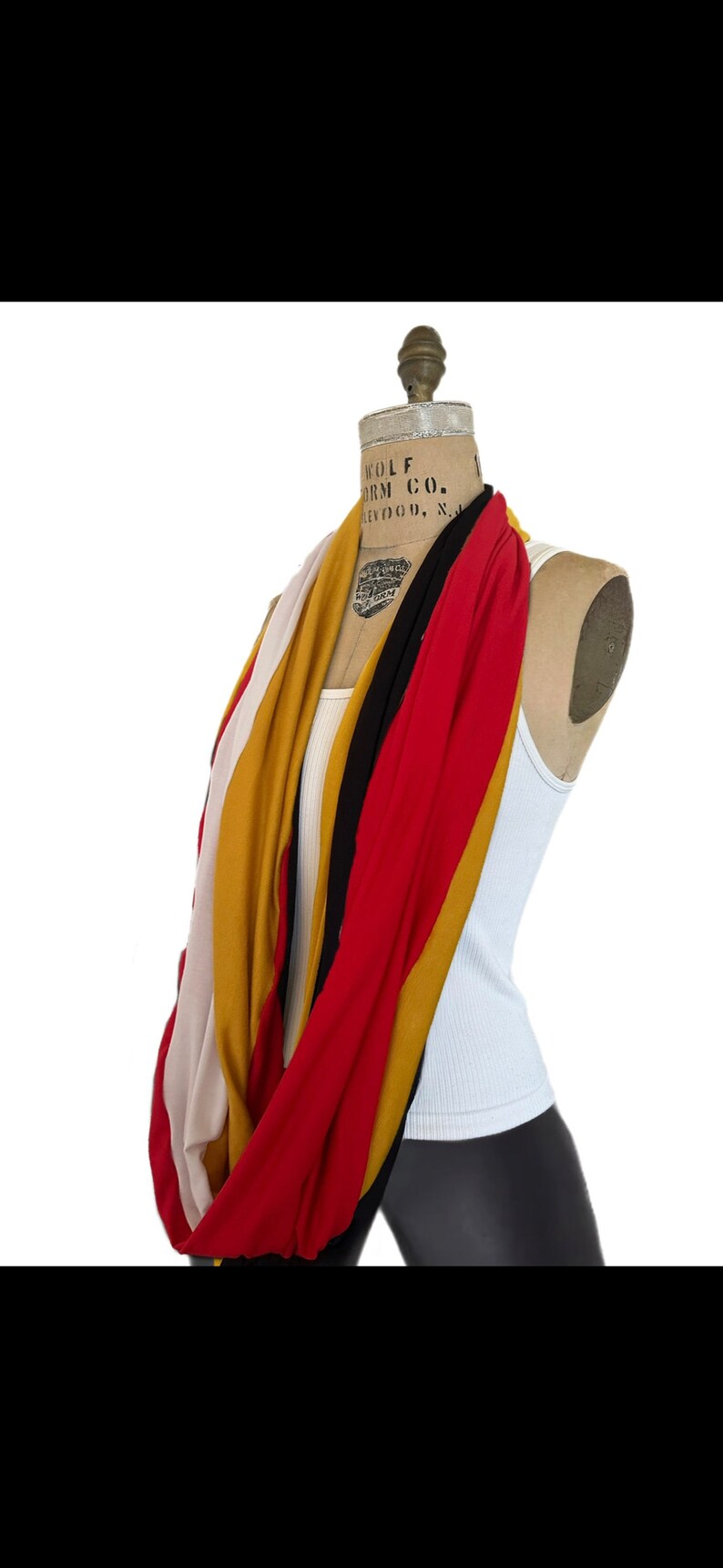 K&ouml;nnte beinhalten: Ein mehrfarbiger Loop-Schal, der auf einer Schaufensterpuppe drapiert ist. Der Schal hat Streifen in Rot, Gold, Schwarz, Creme und Senfgelb. Die Schaufensterpuppe tr&auml;gt ein wei&szlig;es Tanktop und schwarze Leggings. Der Hintergrund ist wei&szlig;.