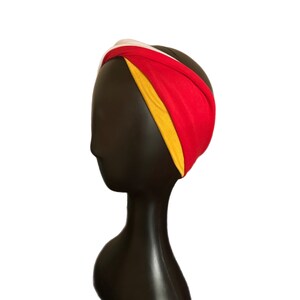 K&ouml;nnte beinhalten: Ein farbenfrohes Stirnband mit einem gedrehten Design. Das Stirnband besteht aus einer Kombination aus rotem, gelbem und wei&szlig;em Stoff. Das Accessoire wird auf einem schwarzen Schaufensterkopf pr&auml;sentiert, um seine Form und seinen Stil hervorzuheben. Dieser Artikel ist ein Modeaccessoire.