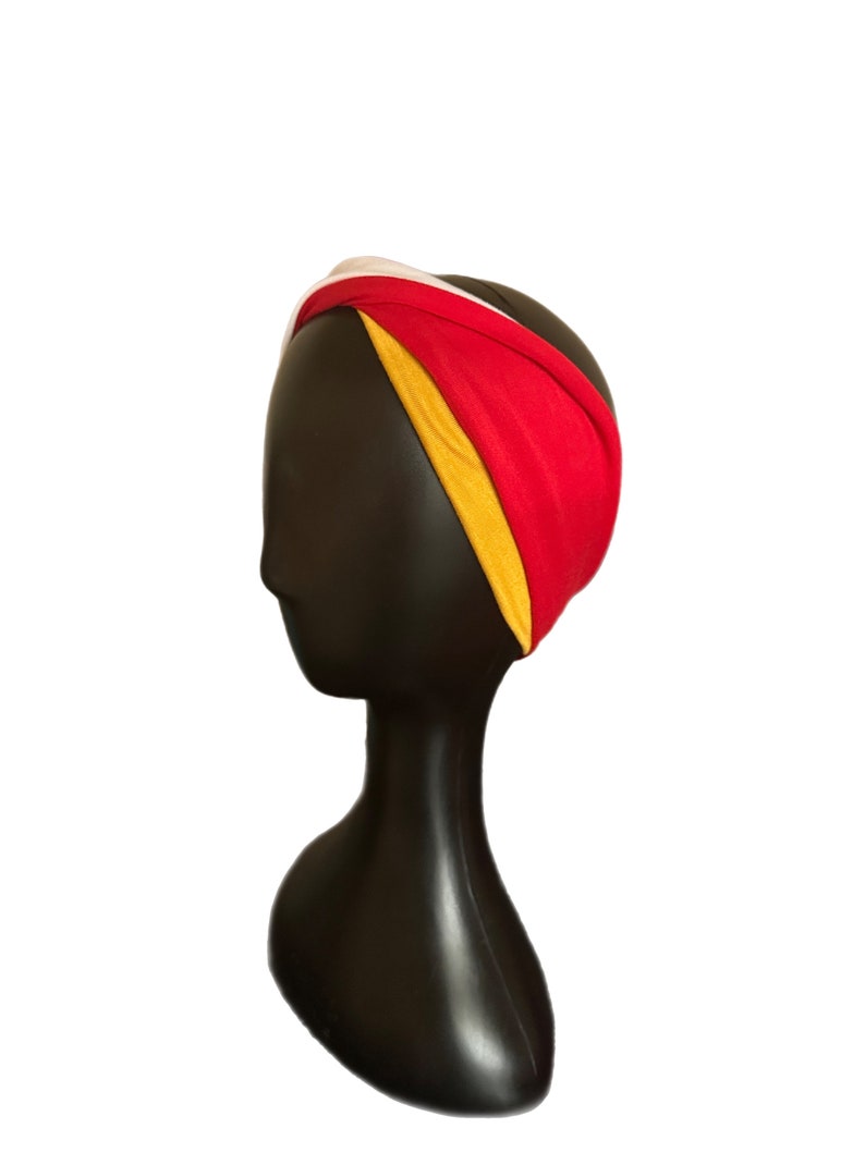 K&ouml;nnte beinhalten: Ein farbenfrohes Stirnband mit einem gedrehten Design. Das Stirnband besteht aus einer Kombination aus rotem, gelbem und wei&szlig;em Stoff. Das Accessoire wird auf einem schwarzen Schaufensterkopf pr&auml;sentiert, um seine Form und seinen Stil hervorzuheben. Dieser Artikel ist ein Modeaccessoire.