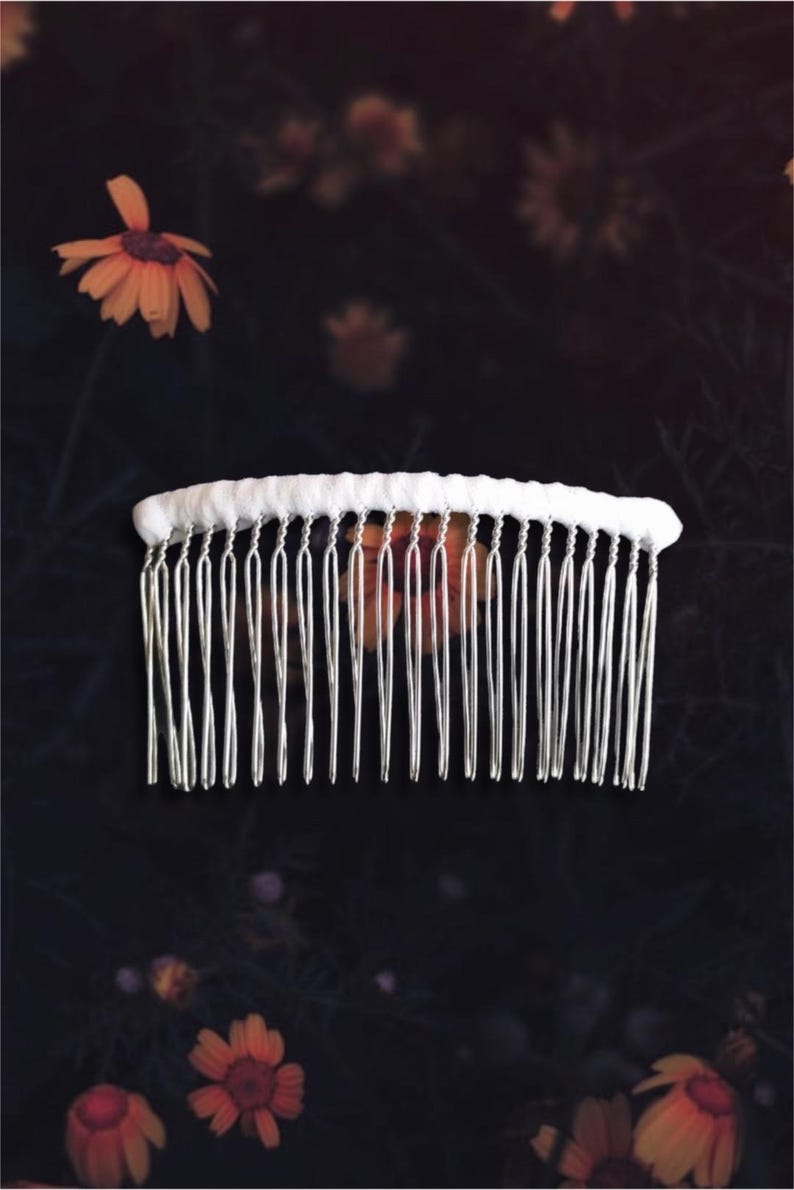 May include: A white fabric-covered hair comb with silver-colored teeth. The comb is set against a dark background with blurred orange flowers. The comb is a hair accessory.