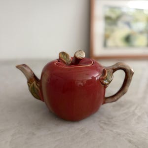 Può includere: Una teiera in ceramica rossa lucida a forma di mela. La teiera presenta un manico a forma di stelo marrone sul coperchio, un beccuccio curvo con dettagli di foglie verdi e un manico laterale marrone a forma di ramo, con finitura smaltata.