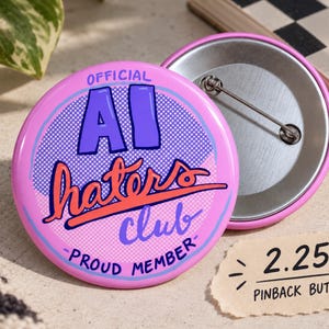 May include: A 2.25-inch round pinback button with a pink background. The design features the text "OFFICIAL AI haters club PROUD MEMBER" in blue and orange. The front shows a purple dotted circle pattern, while the back displays a metal pin mechanism.