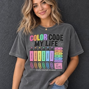 Puede incluir: Una camiseta gris carbón de manga corta con un estampado gráfico que incluye el texto COLOR CODE MY LIFE. El diseño muestra seis resaltadores coloridos con frases, una lista de verificación con iconos, clips de papel y el texto chapter one back to school.