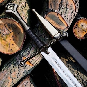 May include: A metal sword with a black leather-wrapped grip, a silver crossguard, and a pommel featuring triangular gold accents. The blade displays an engraved star symbol and runic text. A black scabbard with silver metal fittings lies alongside.