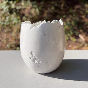 May include: A white, egg-shaped vessel with a smooth, matte finish and a jagged, broken-edge rim. The surface features small, irregular indentations and pits, giving it a textured appearance that resembles a cracked, hollowed-out shell.