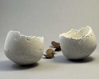 Two small light-gray concrete vessel sculptures. Sustainable. Live edge. Rounded base bowls (wobbly). Shiny outside surface. Wabi-sabi.