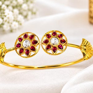 Peut inclure: Un bracelet ouvert en or doté de deux motifs floraux circulaires aux extrémités. Chaque motif contient une pierre ronde transparente centrale entourée de huit pierres rouges en forme de goutte. Les extrémités sont ornées de détails en éventail.