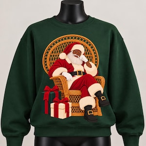 May include: A dark green crewneck sweatshirt featuring a graphic of Santa Claus with a white beard, wearing a red suit with white trim, sitting in a tan wicker peacock chair. Two wrapped gift boxes with red bows are placed at the base of the chair.