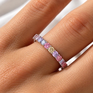 Peut inclure: Une bague alliance en argent ornée d'une rangée de pierres précieuses rondes aux couleurs pastel variées, notamment rose, lavande, bleu clair et jaune pâle. Les pierres sont serties dans une monture à griffes partagées, formant un cercle continu.