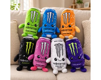 May include: A collection of seven plush toys shaped like Monster Energy drink cans in various colors including green, blue, orange, black, pink, white, and purple. Each plush features a smiling face, small arms and legs, and the Monster logo.