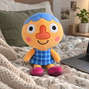 May include: A plush toy featuring a round orange head with blue hair, a prominent orange nose, and large white eyes with black pupils. The character has pink cheeks and a black smile, wearing a blue plaid dress and dark pink shoes.