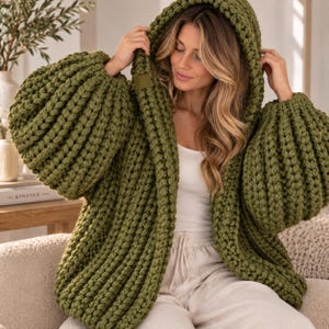May include: An olive green chunky knit cardigan featuring a hood, long voluminous sleeves, and a thick ribbed texture. The garment is crafted from heavy yarn with a prominent stitch pattern, showcasing a relaxed fit and a small rectangular tag.