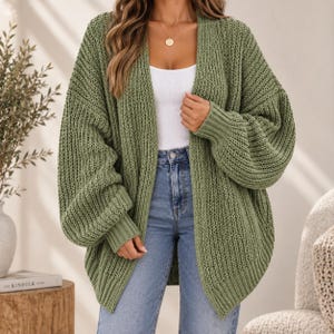 Könnte beinhalten: Eine olivgrüne, offene Strickjacke mit grober Strickstruktur, langen Ballonärmeln mit gerippten Bündchen und einem gerippten Saum. Das Kleidungsstück wird über einem weißen Tanktop und einer blauen Jeans getragen und hat eine lockere Passform.