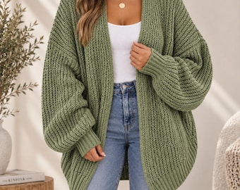 Può includere: Un cardigan aperto color verde oliva con una trama a maglia grossa, maniche lunghe a palloncino con polsini a coste e orlo a coste. Il capo è indossato sopra una canotta bianca e jeans blu, caratterizzato da una vestibilità ampia.