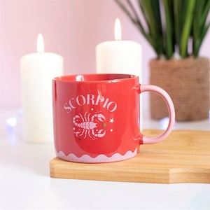 May include: A red ceramic mug featuring the word SCORPIO in white letters above a white scorpion illustration and small star accents. The mug has a light pink handle and a scalloped light pink base design, providing a colorful drinkware option.