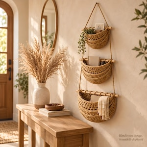 Könnte beinhalten: Ein dreistufiges Wandregal mit geflochtenen beigen Körben, die an Seilen an Holzstäben hängen. Die Körbe enthalten eine kleine Pflanze, Bücher und ein gefaltetes Tuch. Der Text "Handwoven storage, beautifully displayed" ist auf dem Bild zu sehen.