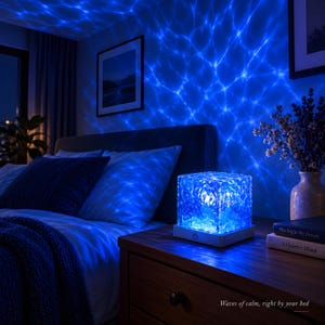 Könnte beinhalten: Eine quadratische Lampe mit kristalliner Textur, weißem Sockel und Ein-Aus-Schalter, die ein hellblaues Licht ausstrahlt und ein wellenförmiges Wassermuster an die Wände projiziert. Der Text "Waves of calm, right by your bed" ist sichtbar.