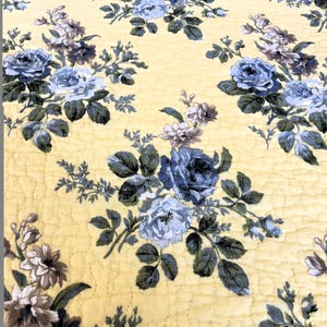 May include: A yellow floral quilt featuring a repeating pattern of blue and gray roses with green leaves. The quilt has a quilted texture with a diamond pattern. The overall design is a classic floral pattern.
