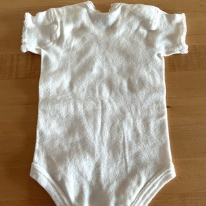 Puede incluir: Body de beb&eacute; blanco con mangas cortas. El body est&aacute; hecho de una tela suave y texturizada. La prenda tiene un dise&ntilde;o sencillo con cuello redondo y corchetes en la parte inferior para facilitar el cambio de pa&ntilde;ales.