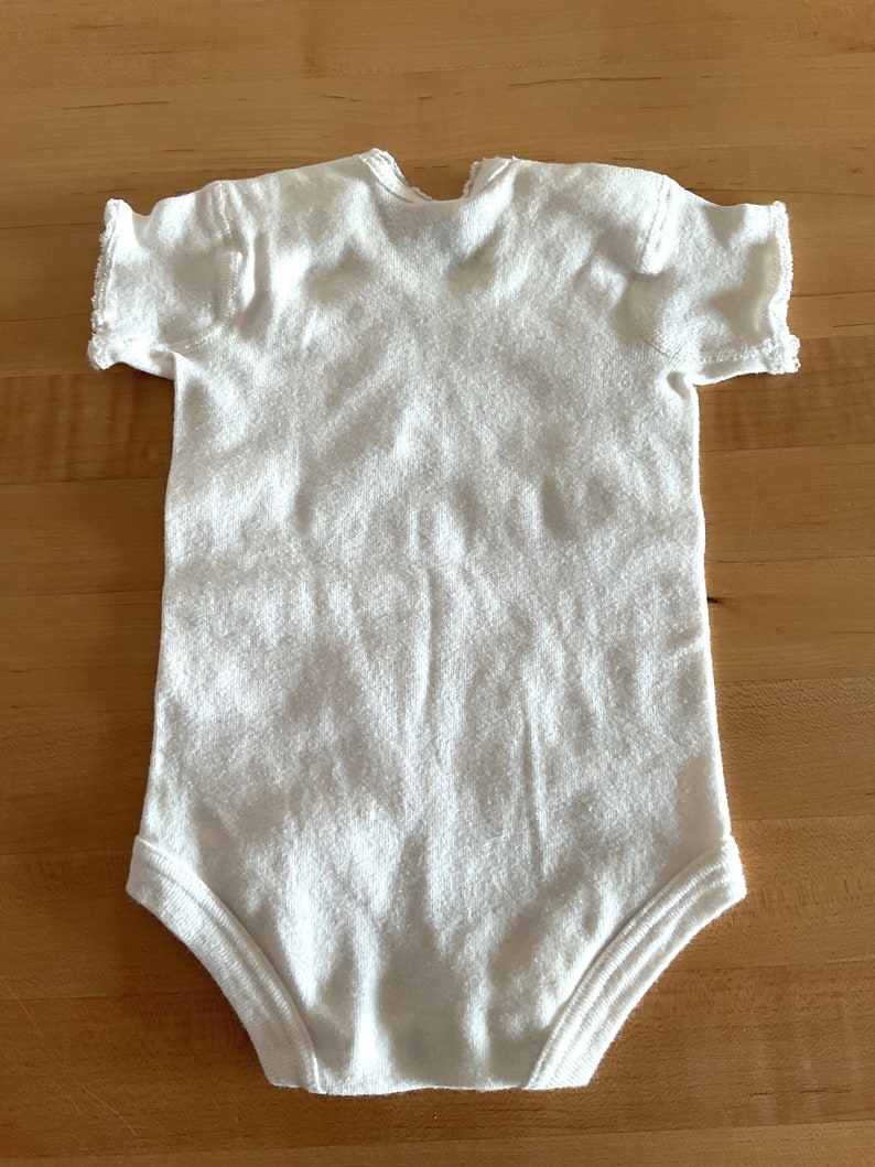 Puede incluir: Body de beb&eacute; blanco con mangas cortas. El body est&aacute; hecho de una tela suave y texturizada. La prenda tiene un dise&ntilde;o sencillo con cuello redondo y corchetes en la parte inferior para facilitar el cambio de pa&ntilde;ales.