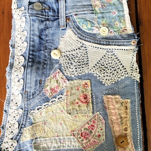 May include: A blue denim fabric piece decorated with white lace trim, floral fabric patches, and buttons. The denim features a metal rivet and a vertical strip of fabric with the word VINTAGE printed on it. The edges are frayed and stitched.