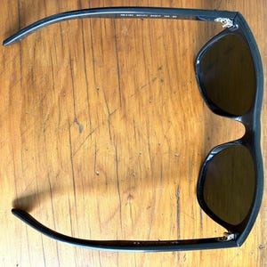 Puede incluir: Gafas de sol negras con lentes oscuras. Las gafas tienen una montura rectangular. Las patillas de las gafas son curvas y se extienden hacia afuera. Sobre un fondo de madera.